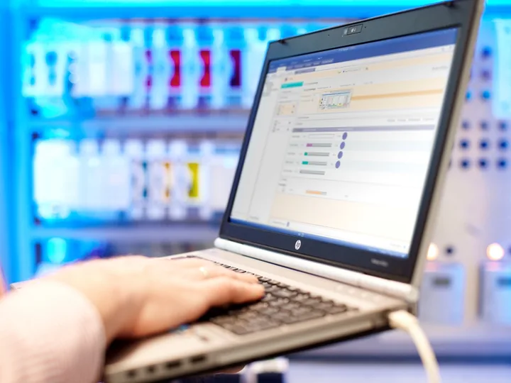Hand auf der Tastatur eines Laptops mit Priva-Software, im Hintergrund ein beleuchteter Schaltschrank mit Automationsmodulen