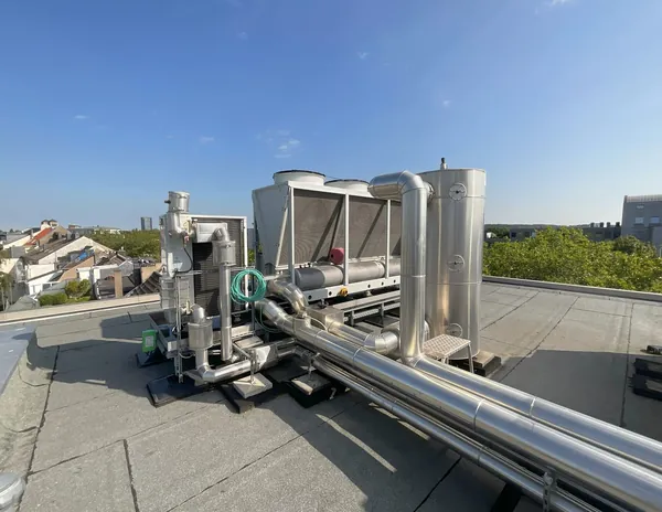 Dachaufstellung einer Kälteanlage: luftgekühlter Kälteerzeuger, Edelstahl-Speicherbehälter und gedämmte Rohrleitungen auf einem Flachdach, dahinter blauer Himmel und die Stadt