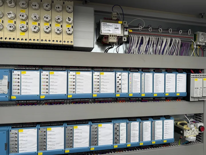Geöffneter Bestandsschaltschrank mit zwei Reihen DDC-Modulen einer älteren Generation, jedes Modul mit Beschriftungsfeld, Handschaltern und Status-LEDs, darüber Sicherungen, Transformator und Klemmleiste
